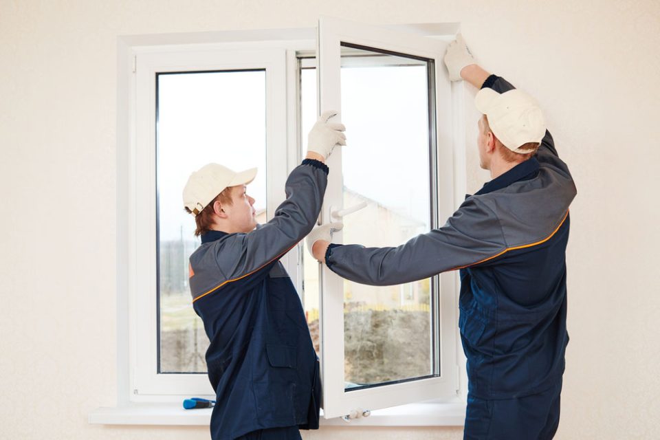 Two construction workers in blue overalls and caps install a white window, guiding the sash outward.