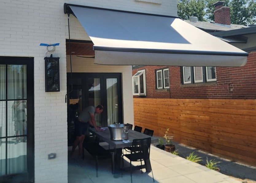 Backyard patio with a large beige retractable awning extended over an outdoor dining table; a man stands and leans over the table near a large pot.